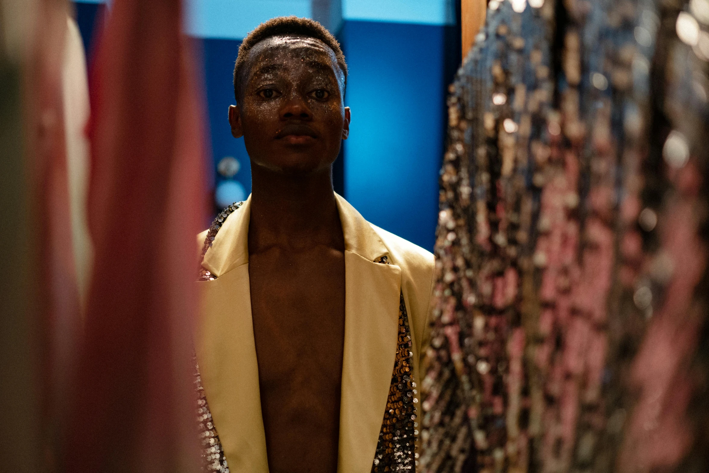 a man standing in front of a rack of clothes, by Daniel Lieske, pexels contest winner, maximalism, brown skinned, sequins, blue gold suit, non binary model