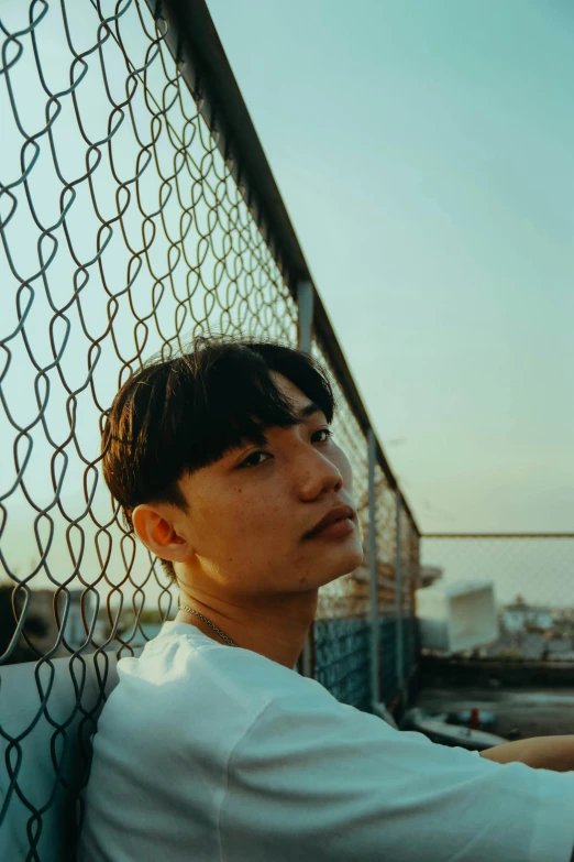 a man sitting on a bench next to a fence, an album cover, inspired by jeonseok lee, unsplash, realism, handsome face, golden hour, rooftop, south east asian with round face