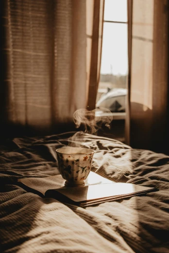 a coffee cup sitting on top of a bed next to a window, pexels contest winner, smoke, sun down, tea cup, gif