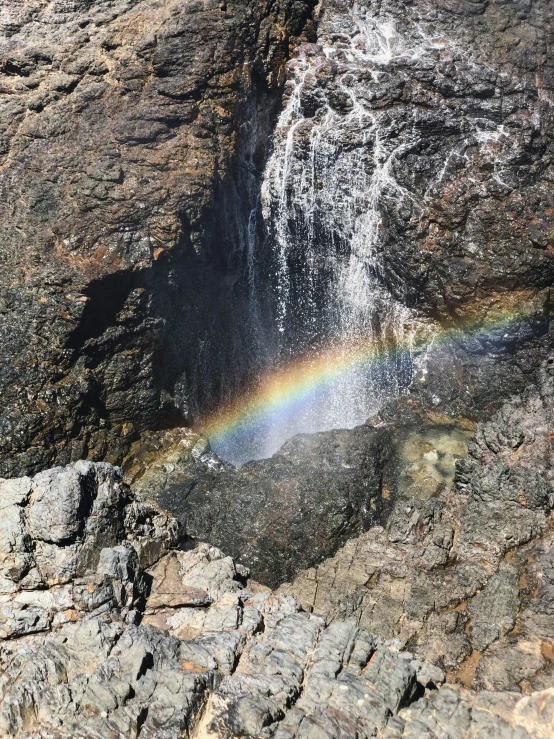 a rainbow is in the middle of a waterfall, with lots of dark grey rocks, sparkling in the sunlight, pareidolia, fully functional