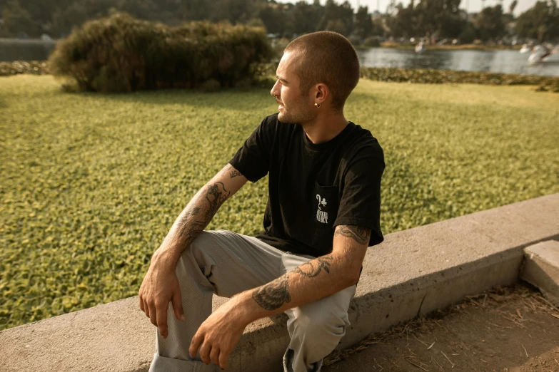 a man sitting on a ledge next to a body of water, wearing black tshirt, federation clothing, in the park, plain walls |light hearted