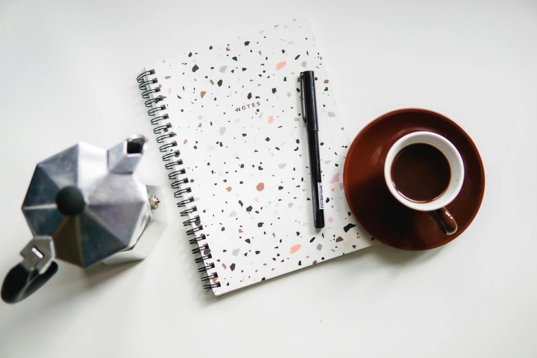 a cup of coffee and a notebook on a table, by Julia Pishtar, pexels contest winner, minimalism, terrazzo, 9 9 designs, white with chocolate brown spots, rpg item