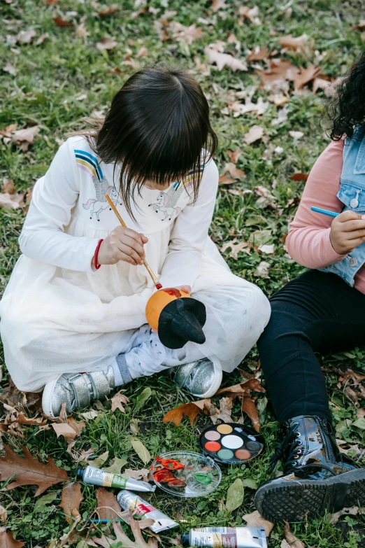 a couple of young girls sitting on top of a grass covered field, a child's drawing, inspired by Diego Velázquez, pexels contest winner, holding a paintbrush, autum, children playing with pogs, black haired girl wearing hoodie