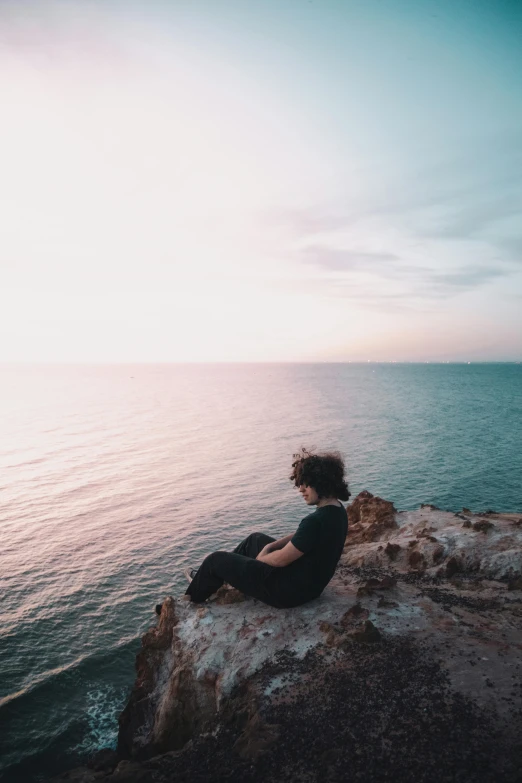a person sitting on top of a rock next to the ocean, in the evening