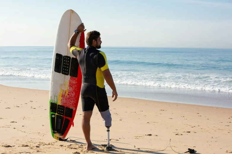 a man standing on a beach holding a surfboard, prostheses, injured, rocha, accessible for the disabled