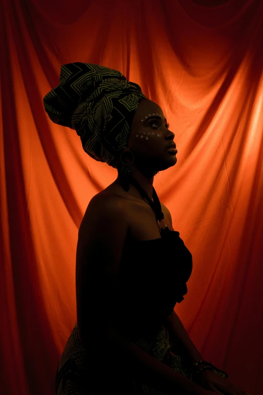 a woman sitting in front of a red curtain, pexels contest winner, afrofuturism, cloth head wrap, dramatic lowkey studio lighting, orange and black tones, black silhouette