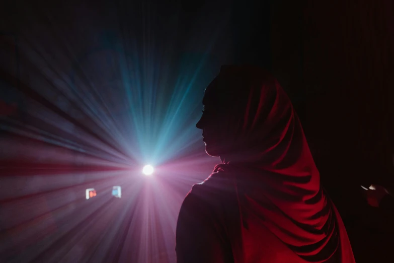 a person standing in front of a bright light, a hologram, by Adam Marczyński, unsplash contest winner, wearing a red cape, an arab standing watching over, she has a glow coming from her, scene from a rave