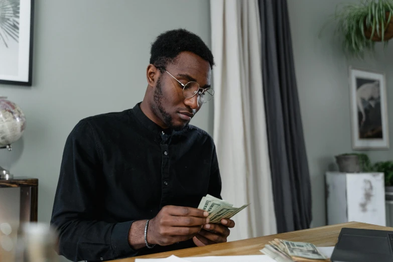 a man sitting at a table with money in front of him, by Meredith Dillman, pexels contest winner, hurufiyya, wearing a black shirt, black people, student, 1 5 0 4