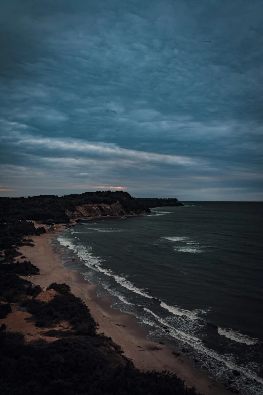 a beach next to a body of water under a cloudy sky, unsplash contest winner, australian tonalism, cliff side at dusk, drone footage, vibrant but dreary blue, looking threatening