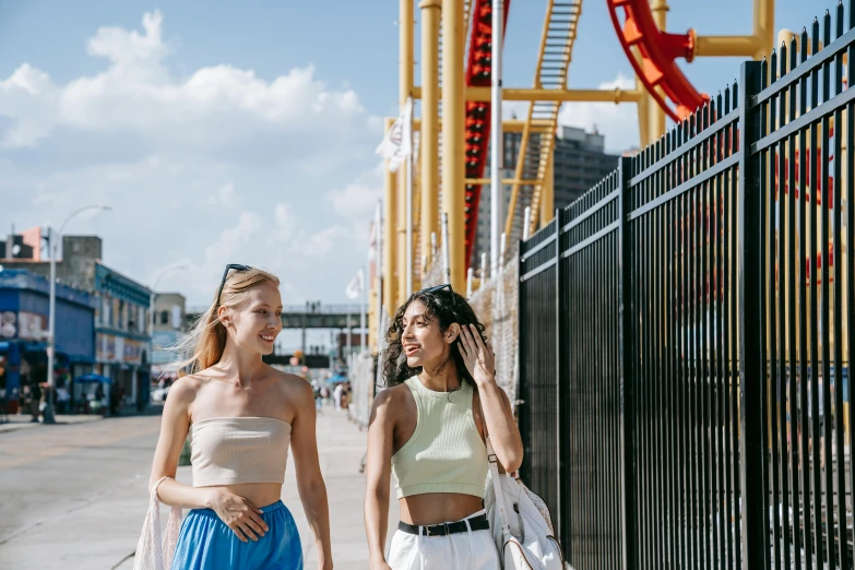 two women walking down a sidewalk next to a roller coaster, trending on pexels, wearing yellow croptop, beaches, brooklyn background, background image