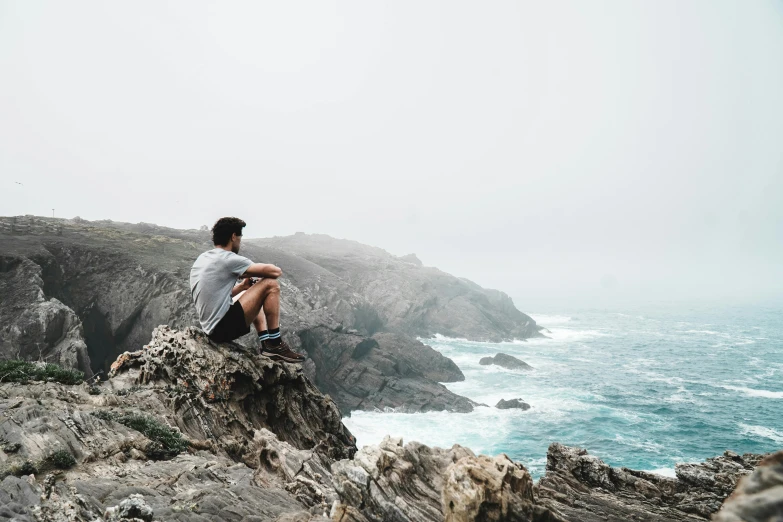 a man sitting on a rock overlooking the ocean, pexels contest winner, gray men, craggy, wondering about others, minimalistic