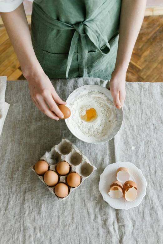 a person that is holding an egg in a bowl, baking a cake, curated collections, ingredients on the table, linen