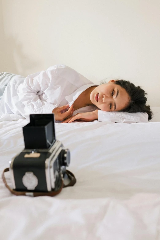 a woman laying on top of a bed next to a camera, a picture, looking tired, hasselblad photograph, college, white