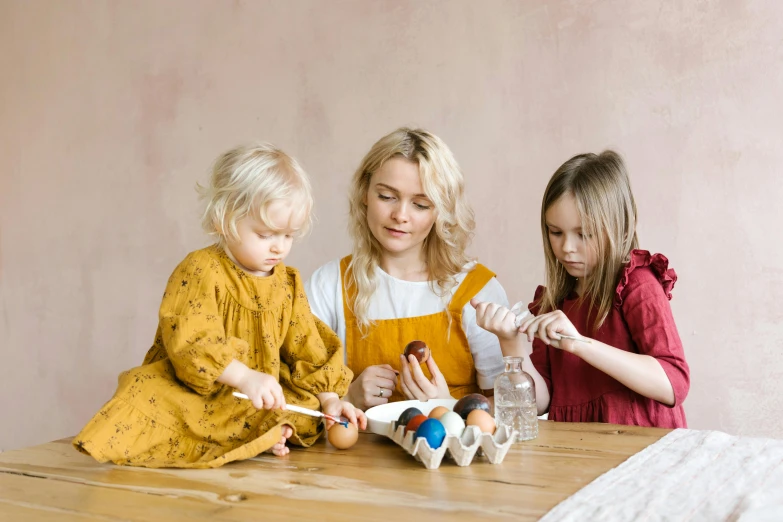 a woman and two children sitting at a table, a still life, inspired by Elsa Beskow, pexels contest winner, eggs, wooden art toys, 15081959 21121991 01012000 4k, wearing an apron
