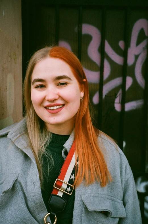 a woman with red hair standing in front of a building, smiling for the camera, pokimane, orange and black tones, non-binary