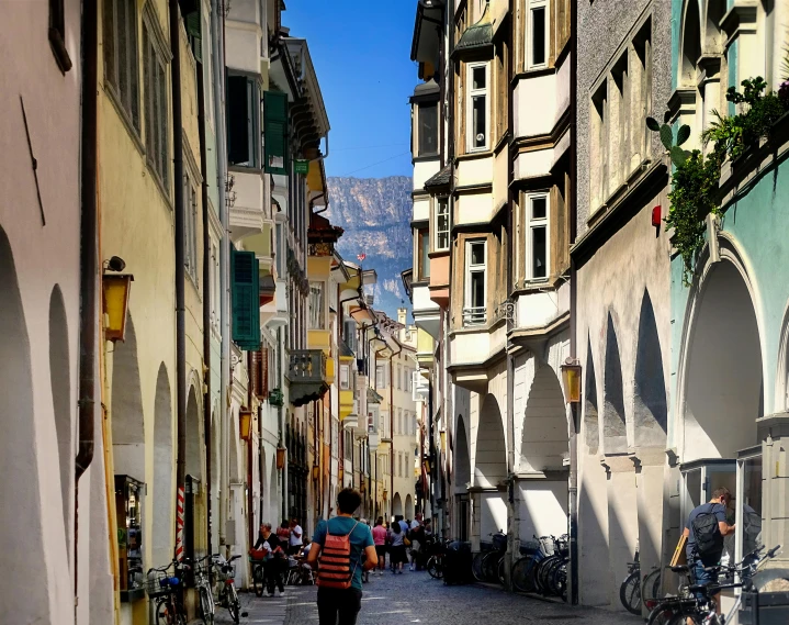 a person walking down a narrow city street, inspired by Karl Stauffer-Bern, pexels contest winner, renaissance, dolomites in background, square, colorful scene, palermo city street