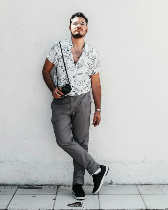 a man leaning against a wall with a camera, by Robbie Trevino, patterned clothing, white grey color palette, promo image, full-body-shot