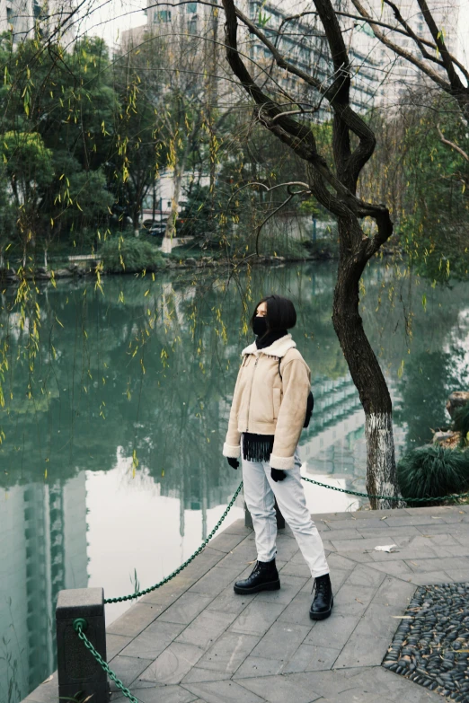 a man standing next to a body of water, an album cover, inspired by Zhang Xuan, trending on unsplash, city park, face covered, wearing jacket, offwhite