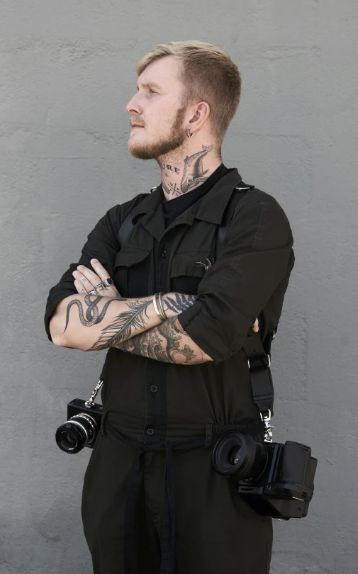 a man with tattoos standing in front of a wall, inspired by Cam Sykes, trending on pexels, tactical vests and holsters, holding a camera, professional profile photo, casual black clothing