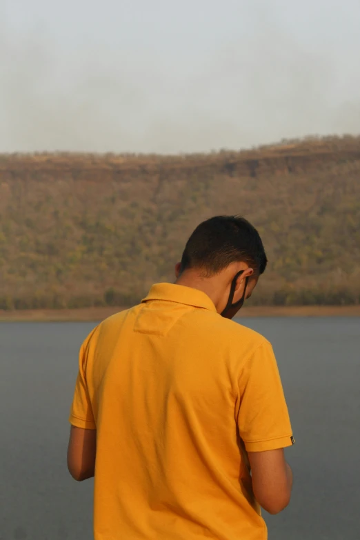 a man standing next to a body of water, by Saurabh Jethani, wearing a modern yellow tshirt, forest fires in the distance, crater, wearing polo shirt