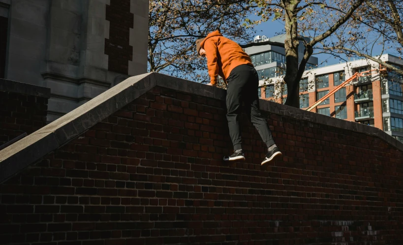 a man riding a skateboard up the side of a brick wall, by Sam Charles, pexels contest winner, happening, orange hoodie, parkour, background image, athletic crossfit build