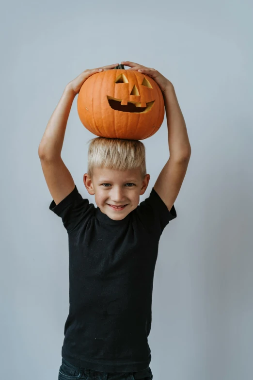 a young boy balancing a pumpkin on his head, pexels contest winner, on a gray background, blond boy, dark. no text, promotional image