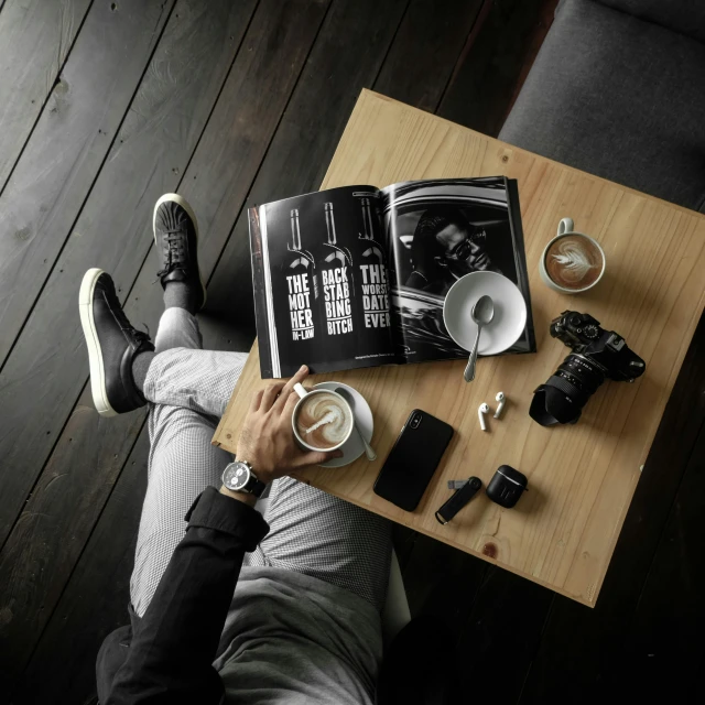 a person sitting at a table with a book and a cup of coffee, by Matthias Stom, pexels contest winner, sneaker photo, hasselblad medium format, all black matte product, bottles of alcohol next to him