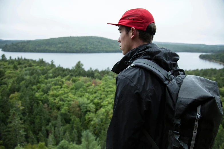 a man with a backpack looking out over a lake, by Jessie Algie, red hat, grey forest in the background, shot from roofline, avatar image