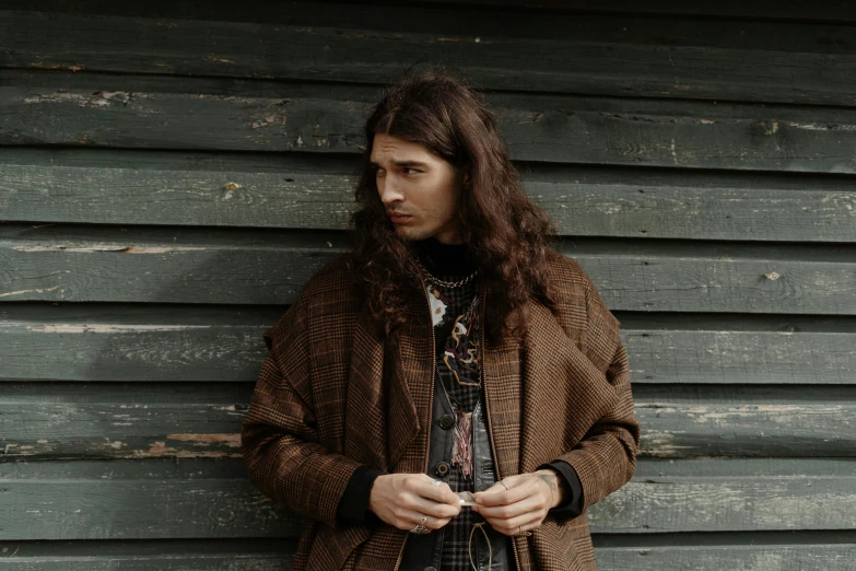 a man standing in front of a wooden wall, an album cover, by Winona Nelson, trending on pexels, renaissance, brown long hair, wearing hunter coat, patterned clothing, hozier