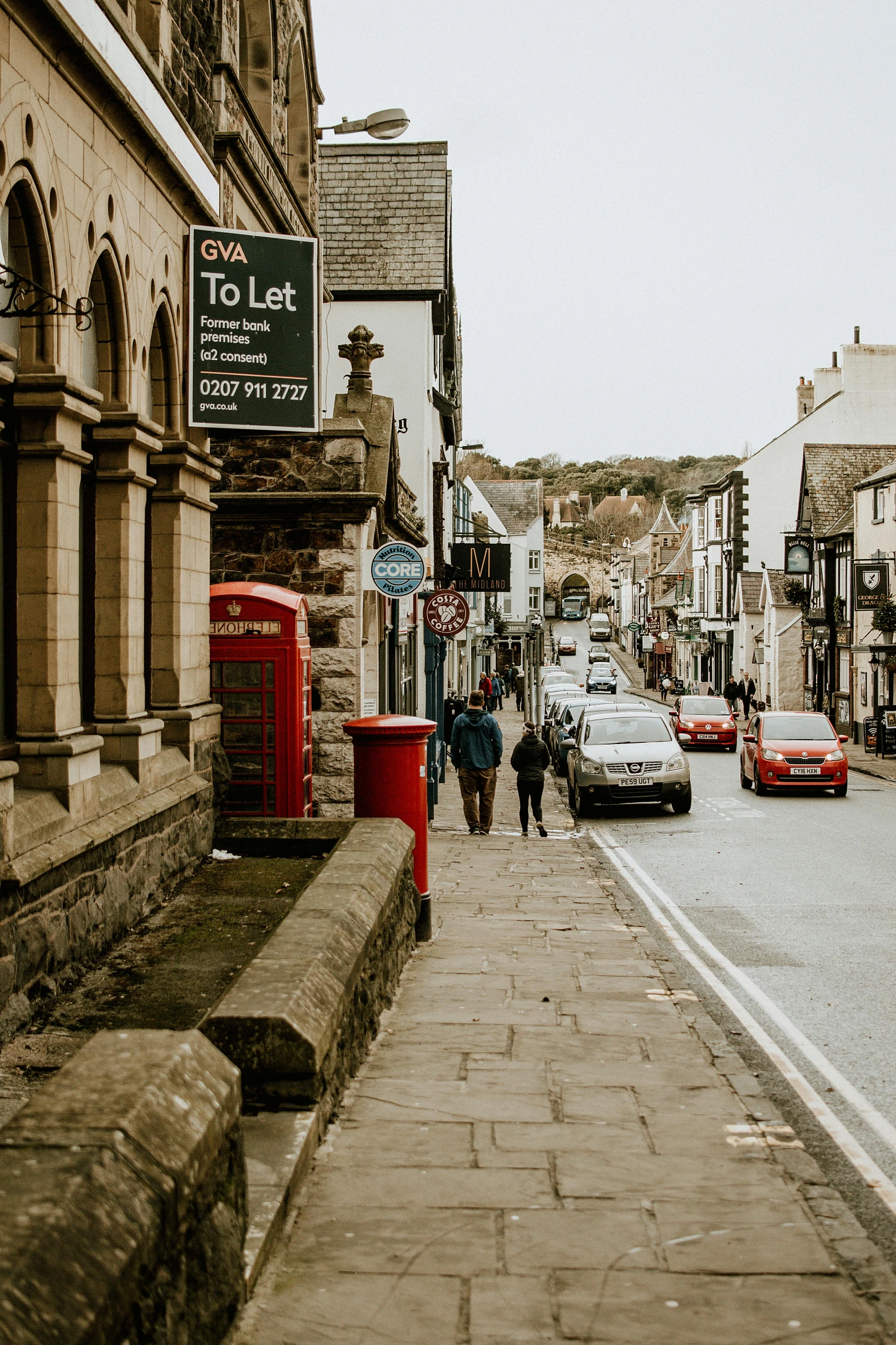 a couple of people walking down a street, by Kev Walker, pexels contest winner, medieval coastal village, letterbox, car, hills