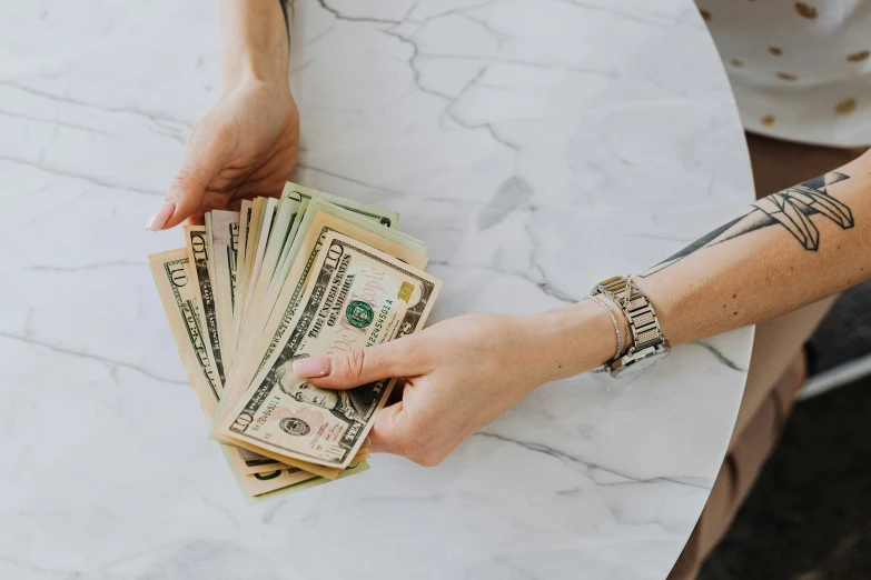 a person holding a stack of money on top of a table, by Emma Andijewska, pexels contest winner, 🦩🪐🐞👩🏻🦳, barbara canepa, loosely cropped, meg kimura