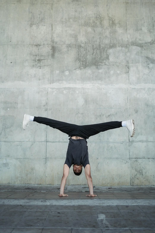 a man doing a handstand in front of a concrete wall, by Nina Hamnett, unsplash, arabesque, plain background, baggy, headshot, large wingspan