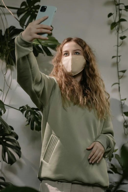 a woman wearing a face mask taking a selfie, inspired by Elsa Bleda, renaissance, greenery, beige, monochromatic, sweater