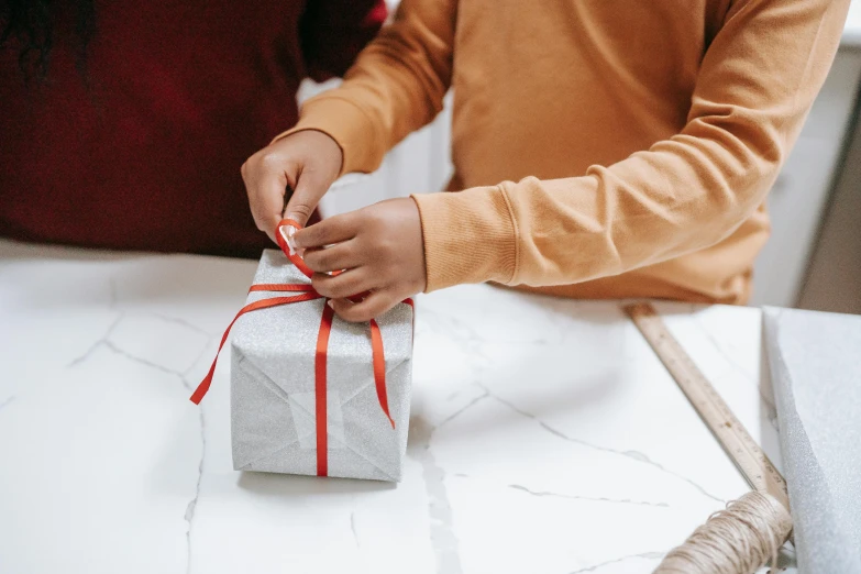 a close up of a person wrapping a present on a table, by Helen Stevenson, pexels contest winner, happening, grey and silver, kids playing, red ribbon, bespoke