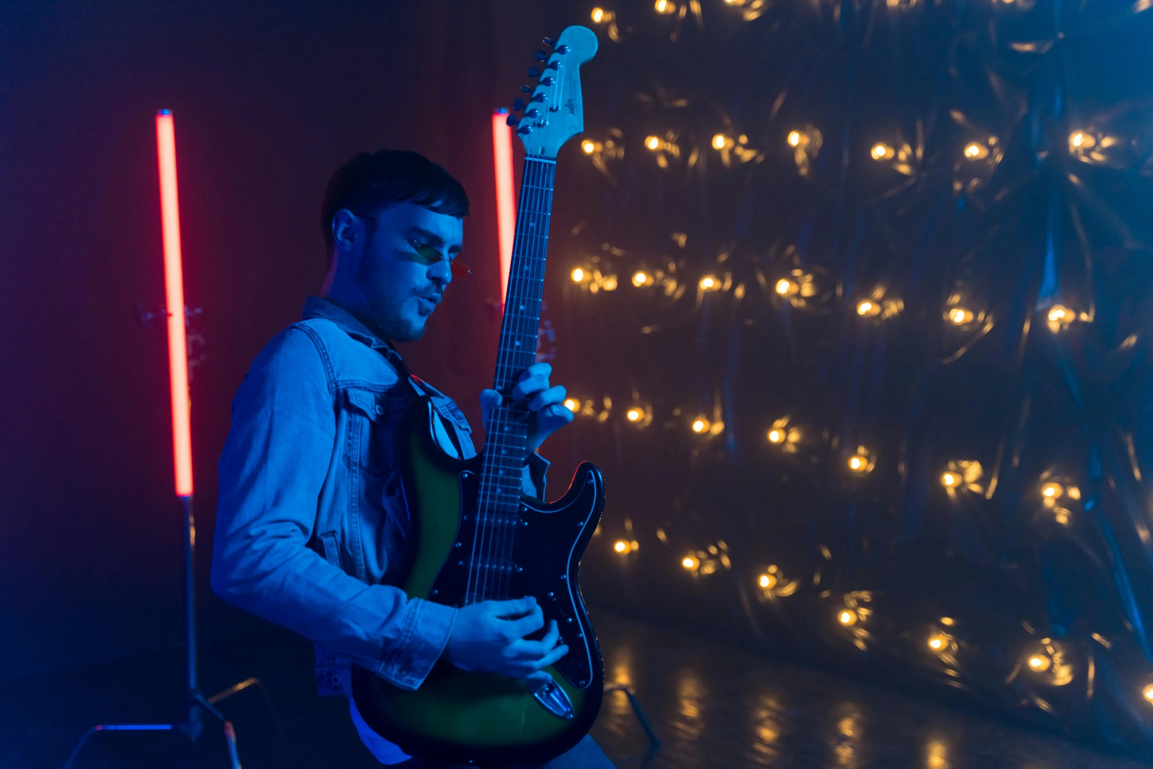 a man sitting on a chair holding a guitar, light and space, neon lights in background, profile image, charli bowater, performing a music video