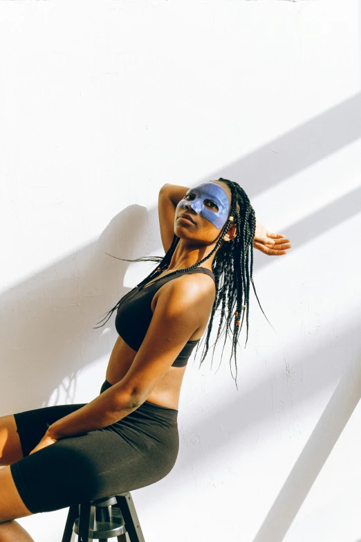 a woman sitting on top of a stool next to a wall, by Nina Hamnett, trending on pexels, afrofuturism, black eye mask, blue body, spa, synthetic bio skin