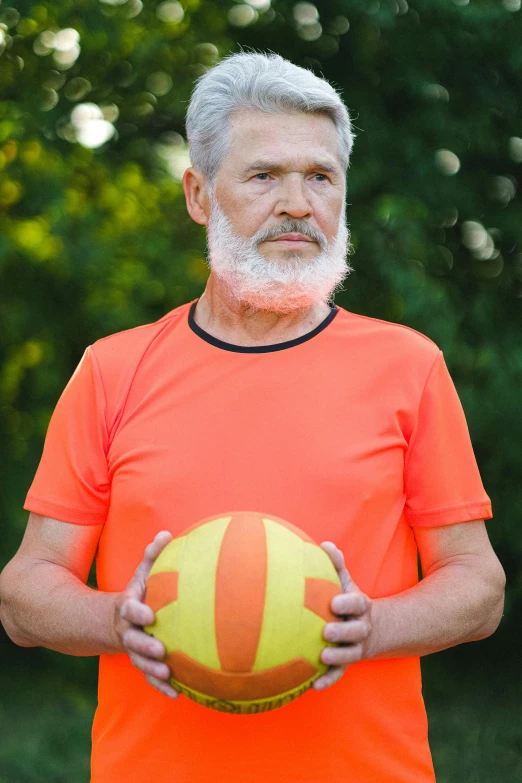 a man in an orange shirt holding a yellow and red ball, bald head and white beard, wearing fitness gear, 2019 trending photo, aging