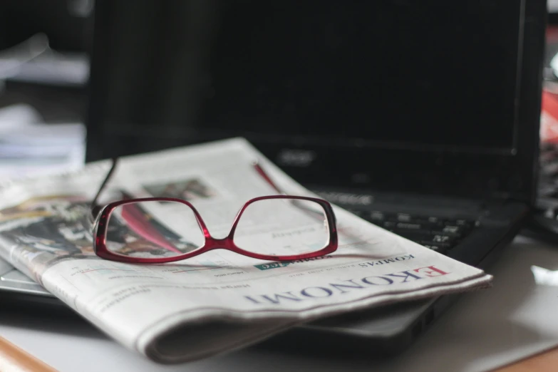 a laptop computer sitting on top of a desk next to a newspaper, by Carey Morris, pexels, square glasses, posing, caucasian, close-up shoot