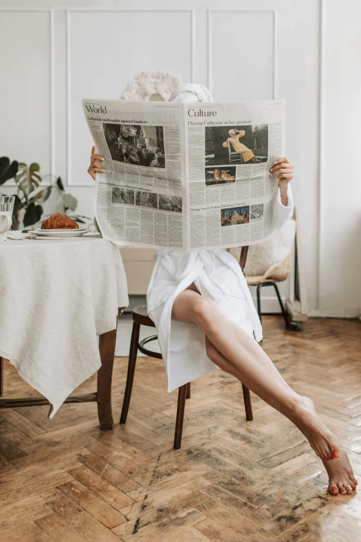 a woman sitting on a chair reading a newspaper, by Maud Naftel, trending on unsplash, private press, white tablecloth, doing a majestic pose, good housekeeping, white robe