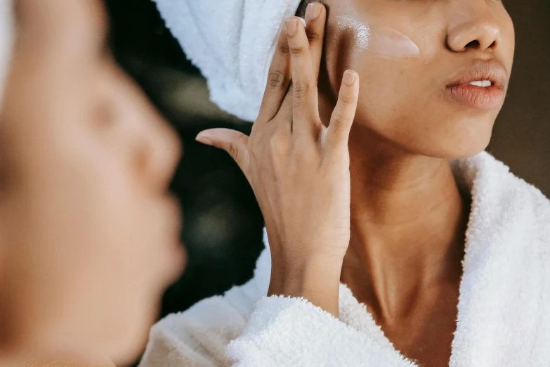 a woman putting cream on her face in front of a mirror, a picture, by Emma Andijewska, trending on pexels, renaissance, square facial structure, covered in robes, manuka, deep tan skin