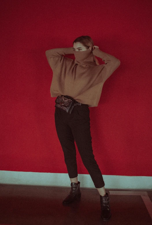 a woman standing in front of a red wall, an album cover, inspired by Elsa Bleda, trending on pexels, renaissance, red sweater and gray pants, wearing all black mempo mask, holster, wearing turtleneck