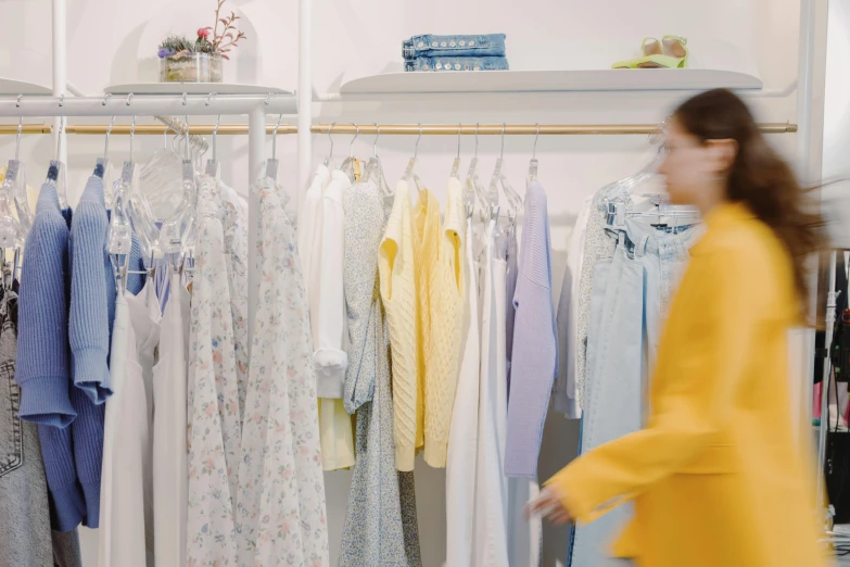 a woman standing in front of a rack of clothes, trending on unsplash, process art, white and yellow scheme, low quality footage, people shopping, wearing a hospital gown