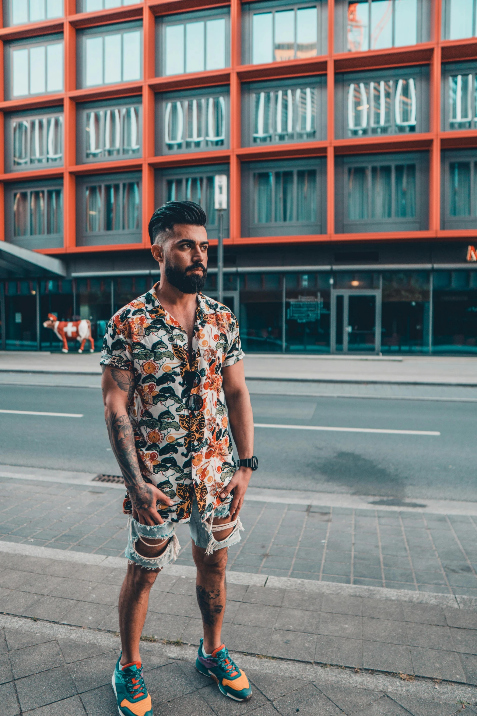 a man standing on a sidewalk in front of a building, by Niko Henrichon, pexels contest winner, body covered in floral tattoos, tan skin a tee shirt and shorts, hannover, zayn malik