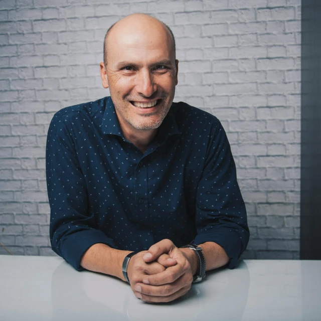 a man sitting at a table in front of a brick wall, a picture, unsplash, no hair completely bald, smiling male, david noren, on grey background