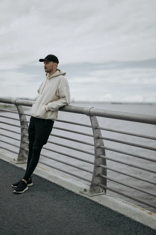 a man standing on top of a bridge next to a body of water, inspired by Jesper Ejsing, unsplash, wearing a track suit, thoughtful pose, standing on street corner, coastal