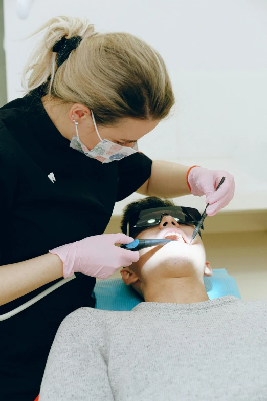 a woman getting her teeth examined by a dentist, by Adam Marczyński, pexels contest winner, square masculine jaw, wearing facemask and sunglasses, high light on the left, glassy fracture