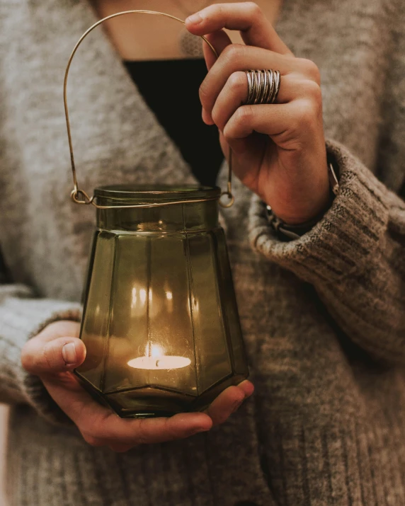 a woman holding a lit candle in her hand, by Helen Stevenson, pexels contest winner, light and space, glass jar, green and warm theme, grey, lantern