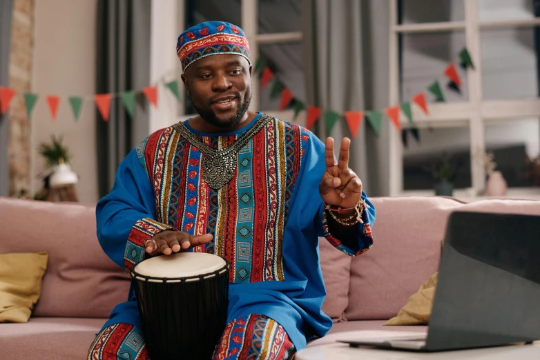 a man sitting on a couch in front of a laptop, an album cover, inspired by Ras Akyem, pexels contest winner, hurufiyya, playing drums, wearing an african dress, youtube thumbnail, congas