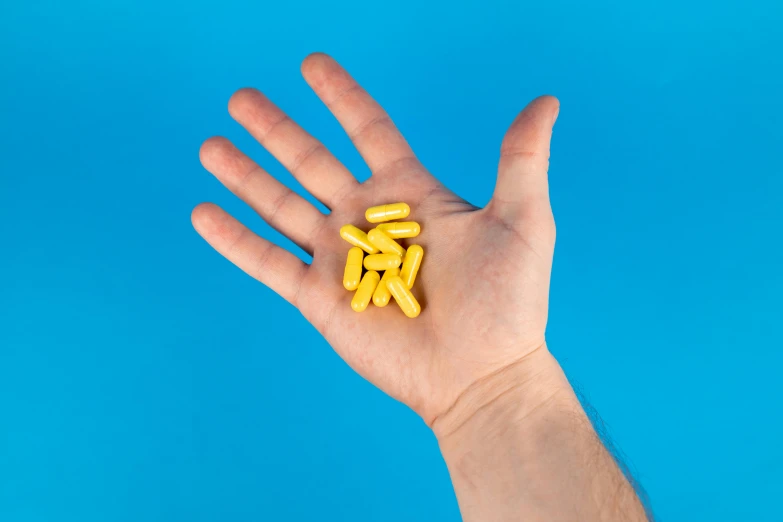 a hand holding yellow pills against a blue background, pexels, taken with sony alpha 9, pose 1 of 1 6, orelsan, made of glazed