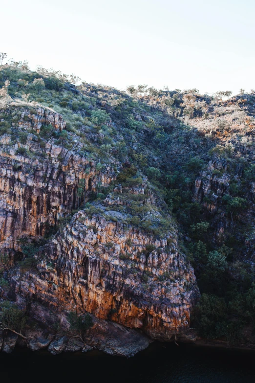 a group of people standing on top of a cliff next to a body of water, australian tonalism, “ aerial view of a mountain, deep crevices of stone, striations, buttresses